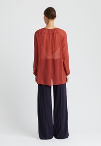 Langes, durchsichtiges, rostfarbenes Blusenshirt mit gerafftem Ausschnitt. Hat lange Ärmel und wird über einer dunkeln, marineblauen, weiten Hose getragen.