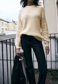 Krémový pletený svetr s texturovanými žebrováním a krajkovými detaily na rukávech, kombinovaný s černými džíny rovného střihu. Šedá bunda držená v ruce.