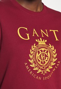 Sudadera burdeos con cuello de canalé que presenta un diseño de logo y escudo en dorado; texto "GANT AMERICAN SPORTSWEAR" en el borde inferior.