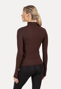 Haut marron à manches longues avec un col montant, design ajusté et texture lisse, associé à des leggings noirs. Les détails des coutures accentuent la silhouette.