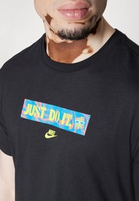Svart bomullströja med rund hals som har en stor flerfärgad grafik med texten "JUST DO IT!" och en liten gul Nike-logotyp längst ner.