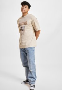 Beige bomulds t-shirt med "AUTHORISERET FORHANDLER" tryk, foto detalje, korte ærmer, parret med lyseblå slidte jeans og sorte sneakers.