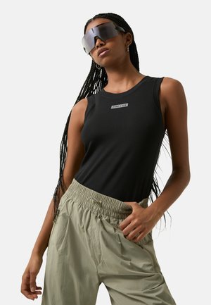 Schwarzes, geripptes Tanktop mit Logodetail, kombiniert mit hellgrünen Cargo-Hosen mit elastischem Bund. Transparente Sonnenbrille, lang geflochtenes Haar.