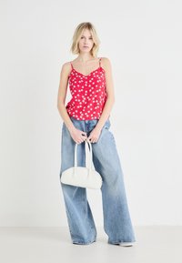 Top in camicia con stampa floreale rossa e scollatura a zig zag, abbinato a jeans oversize in denim blu chiaro. Tiene una piccola borsa bianca.