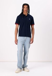 Navy polo shirt with red accents, cotton material, short sleeves, paired with light blue jeans and white sneakers. Simple, fitted design.