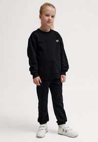 Svart sweatshirt och matchande joggers i mjukt tyg. Sweatshirten har en liten vit logotyp. Modellen bär vita sneakers.
