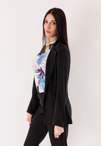 Blazer nero sopra una maglia con motivo multicolore che presenta blu, rosa e giallo. L'outfit ha una silhouette aderente e una texture liscia.