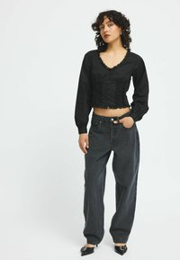 Blusa nera cropped con bordo in pizzo e chiusura a bottoni, abbinata a jeans larghi in denim scuro. La modella indossa scarpe nere a punta.