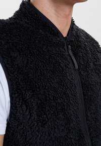 Schwarze Fleece-Weste mit einem Reißverschluss, die eine samtige Textur und einen hohen Kragen aufweist. Das Design umfasst einen einfarbigen schwarzen Saum entlang der Kanten.