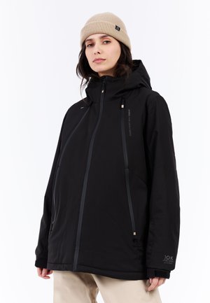 Schwarze wasserdichte Jacke mit Kapuze, mit grauen Akzentreißverschlüssen und Logo. Getragen über beigen Hosen, mit elastischen Bündchen und Reißverschlusstaschen.