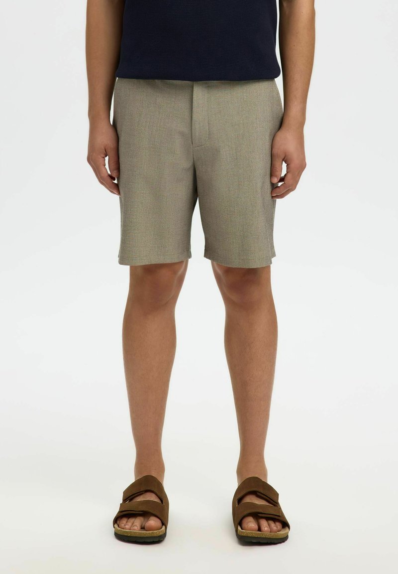 Homme portant un short beige jusqu'aux genoux, un haut noir et des sandales marron à bout ouvert, debout sur un fond blanc.