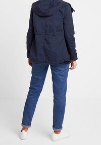 Marineblaue Kapuzenjacke mit Kordelzug in der Taille, strukturierter Stoff und elastische Bündchen, kombiniert mit hochgekrempelten blauen Jeans und weißen Schuhen.