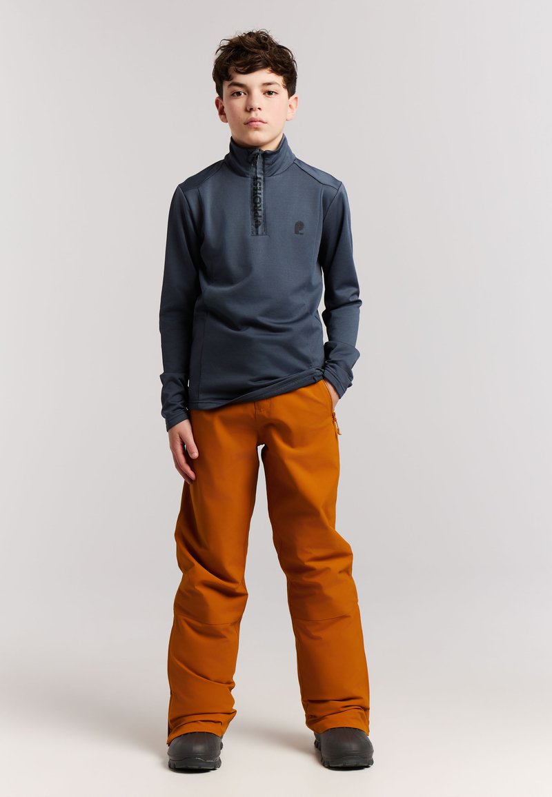Garçon debout portant une veste bleu foncé zippée et un pantalon de neige orange avec des bottes noires sur un fond clair uni.