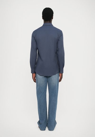 Chemise boutonnée navy à manches longues, avec un ourlet arrondi et des poignets ; associée à un jean large bleu clair, le tout présenté de dos.