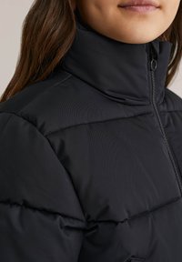 Veste en doudoune noire avec un col haut, dotée d'une fermeture éclair et d'une texture matelassée. Tissu lisse avec des accents de surpiqûres horizontales.