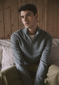 Pull-over zippé gris en tissu doux, avec des manches longues et un col haut, associé à un pantalon vert olive clair. Fond neutre.