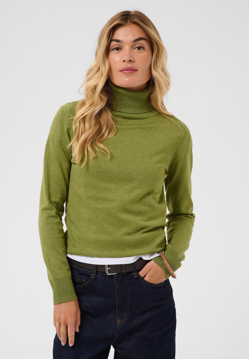 Femme aux longs cheveux blonds portant un pull à col roulé vert olive, un jean foncé et un bracelet en bois à son poignet droit.