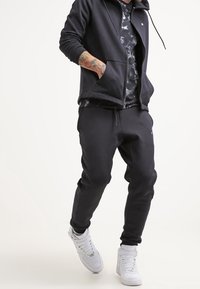 Sudadera con cremallera negra y joggers a juego, hechos de un tejido suave. La sudadera tiene un bolsillo delantero; combinada con zapatillas altas blancas.