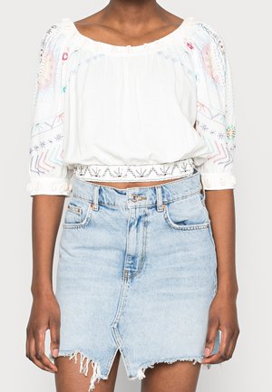 Frau trägt eine weiße bestickte Bluse mit Dreiviertelärmeln und einen hellblauen, ausgefransten Jeansrock mit einem Schlitz vorne.