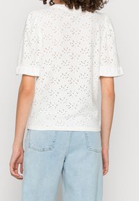 Witte katoenen top met oogjes uitsparingen, korte pofmouwen en een ronde zoom. Gecombineerd met lichtblauwe, hoge taille denim jeans.