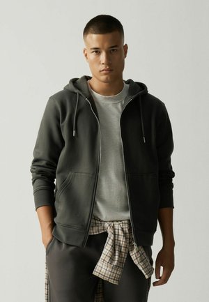 Man draagt een donkerkleurige hoodie met rits over een lichtgrijs T-shirt met een geruit hemd om zijn middel gebonden, staand tegen een effen achtergrond.
