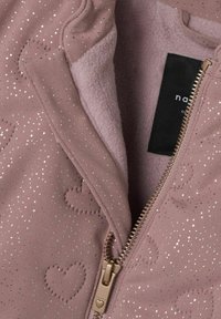 Giacca rosa con motivi a cuore dorati e dettagli glitterati, parzialmente aperta per mostrare l'interno in morbido pile e il tirazip a forma di cuore.