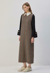 Juoda suknelė su geometriniu raštu, sudarytu iš raudonų, geltonų ir baltų formų, su balta apykakle ir ilgomis rankovėmis su sagų akcentais.