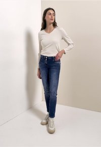 Valkoinen pitkähihainen V-kaula-aukollinen paita, yhdistetty sinisiin skinny-farkkuihin ja beigenvärisiin lenkkarimaisiin kenkiin, näyttäen rennon, istuvan muotoilun.