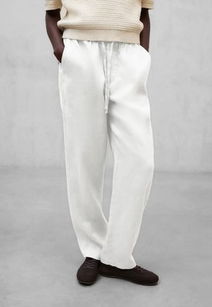 Person iført løstsiddende hvide bukser med snører, beige strikket kortærmet top og sorte sko, stående med hænderne i lommerne på gråt gulv.