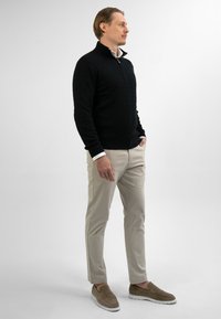 Man in zwart gebreid sweater, beige broek en bruin suède instappers met handen in de zakken op een effen witte achtergrond.