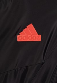 Musta kangas, jossa on punainen kuminen Adidas-logo kolmen viivan kanssa ja sana "adidas" alla, kiinnitetty selkeästi pinnalle.