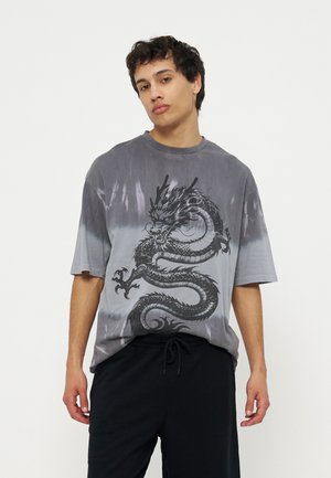 Fiatal férfi szürke és fekete ombre pólót visel, nagy fekete sárkány grafikával, fekete zsinóros nadrággal párosítva.