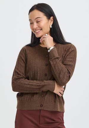 Femme souriant les yeux fermés, portant un cardigan marron à boutons, un pantalon bordeaux et des bijoux en argent sur un fond clair uni.