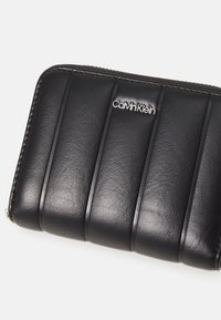 Crni kožni novčanik s vertikalnim šavovima, glatkom teksturom i srebrnim logotipom Calvin Klein na prednjoj strani. Zatvaranje na patentni zatvarač.