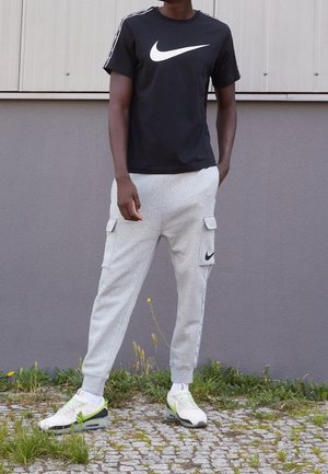 Mężczyzna ubrany w czarną koszulkę Nike, jasnoszare spodnie dresowe typu cargo z czarnym logo Nike oraz białe sneakersy, stojący na brukowanej ścieżce otoczonej trawą.