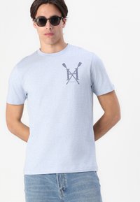 Šviesiai mėlynas medvilninis marškinėliai su trumpomis rankovėmis ir apvaliu iškirpimu. Krūtinėje yra tamsiai pilkas logotipas su kirptais ir užrašu „HACKETT“. 