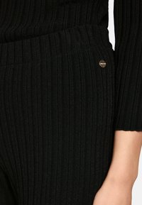 Black ribbed knit top with a fitted shape, featuring a subtle sheen and a small metallic accent on the side. Textured fabric detail visible.