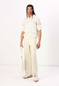 Pull à manches courtes en crème avec des motifs texturés, associé à un pantalon large beige clair et des baskets blanches, tenant un sac fourre-tout crème.