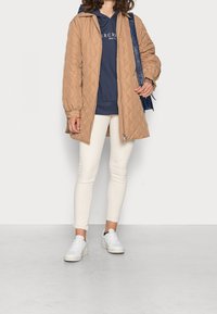 Beżowa, pikowana długa kurtka z zamkiem i kapturem, noszona na ciemnoniebieskim hoodie, w zestawieniu z białymi spodniami i białymi sneakersami.