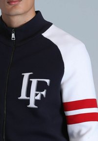 Marineblaues Zip-Jacket mit weißen Raglan-Ärmeln, roten Streifenakzenten und gesticktem "IF"-Logo. Hergestellt aus glattem, strukturiertem Stoff.