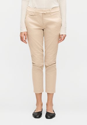 Femme portant un pantalon beige court, un haut blanc à manches longues et des chaussures plates noires, debout devant un fond uni.