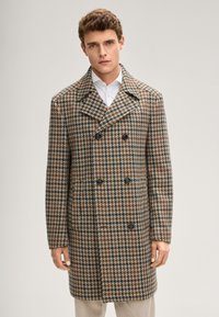 Brauner und beiger Hahnentritt-Mantel mit doppelreihigem Design, großem Kragen und schwarzen Knöpfen, getragen über einem weißen Hemd.