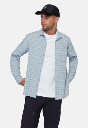 Mann trägt ein hellblau kariertes Hemd mit Knöpfen über einem weißen T-Shirt, schwarze Hosen mit Reißverschlusstasche und eine schwarze Baseballkappe, Hände in den Taschen.