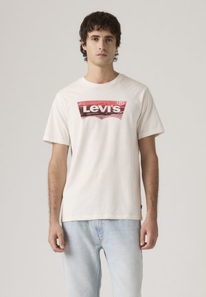 Νέος άνδρας όρθιος, φοράει λευκό t-shirt Levi's με κόκκινο και μαύρο λογότυπο και ανοιχτόχρωμο τζιν, μπροστά σε απλό φόντο.