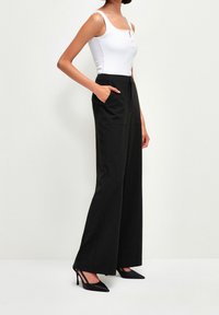 Pantalons larges noirs avec deux poches latérales, coupe cintrée et texture de tissu lisse, associés à un haut blanc ajusté et des talons noirs.