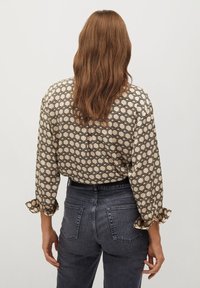 Femme aux longs cheveux bruns portant un chemisier beige à motifs et un jean gris foncé taille haute, debout devant un fond blanc uni.