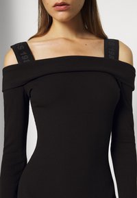 Femme portant une robe noire côtelée à épaules dénudées avec de larges bretelles noires, recadrée de la bouche à la taille.