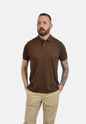 Uomo con barba e tatuaggi che indossa una polo marrone a maniche corte e pantaloni beige, in piedi con le mani nelle tasche davanti a uno sfondo uniforme.