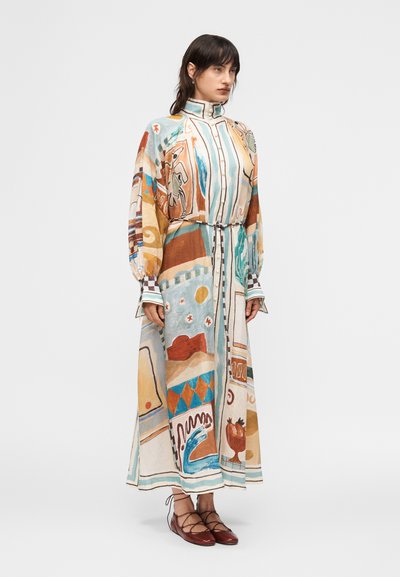 Γυναίκα με μακρύ, πολύχρωμο φόρεμα με αφηρημένα σχέδια, μακριά μανίκια, ψηλό γιακά και λεπτή ζώνη, συνδυασμένο με μπορντό παπούτσια δαντέλας με κορδόνια.
