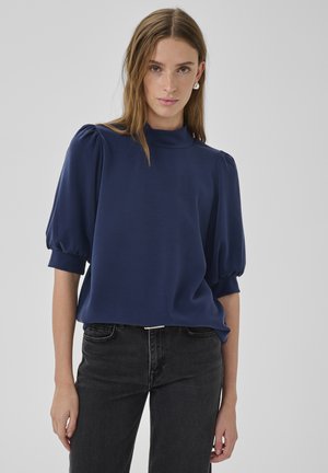 Tânără purtând o bluză bleumarin cu mâneci bufante, guler fals și blugi negri cu talie înaltă, stând pe un fundal gri.